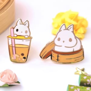 May include: Two enamel pins featuring a white bunny. One pin shows a bunny peeking out of a yellow and brown boba tea cup with a pink straw. The other pin shows a bunny sitting in a yellow and brown steamer basket.