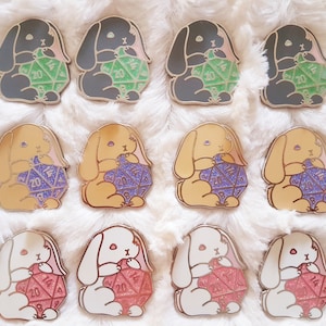 Op de afbeelding: Tien emaillepins met verschillende schattige cartoon konijnen die verschillende gekleurde dobbelstenen vasthouden. De konijnen staan in verschillende poses en hebben verschillende vachtkleuren. De dobbelstenen zijn allemaal verschillend gekleurd en hebben verschillende nummers.