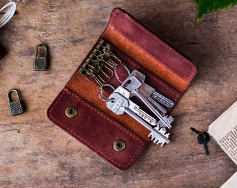 May include: A dark brown leather key case featuring a brass-toned six-hook organizer and two snap button closures. The interior holds several silver-toned metal keys of varying shapes and sizes, secured to the hooks within the folded leather wallet.