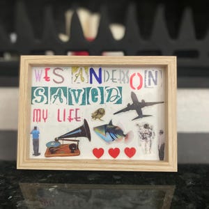 Op de afbeelding: Een ingelijst kunstwerk met de tekst "Wes Anderson Saved My Life" in kleurrijke letters. Het kunstwerk bevat afbeeldingen van een grammofoon, een vis, een vliegtuig, harten en een astronaut. Het frame is gemaakt van lichtgekleurd hout.