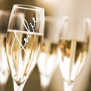 May include: A close-up of a champagne flute with the name "Jodie" written on the side. The flute is filled with champagne and there are other flutes in the background.