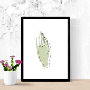 May include: Black framed art print featuring a minimalist line drawing of a hand with a light green fill. The artwork is set against a white background. A small white pot with pink flowers is in the foreground.