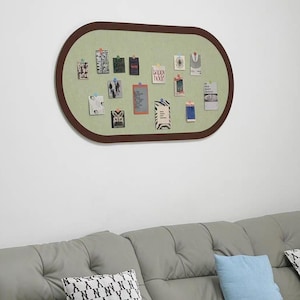 Puede incluir: Un tablón de anuncios ovalado con un interior verde claro y un marco marrón oscuro. Varias tarjetas y notas pequeñas están sujetas al tablero. Un sofá gris con cojines decorativos está en primer plano.