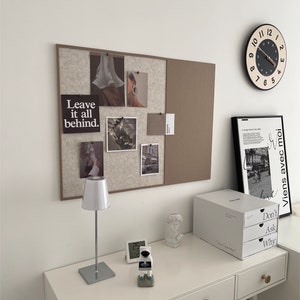May include: A white desk with a white lamp, a clock, and a white storage box with three drawers. The drawers have the words "Don't Ask Why" printed on them. The desk is against a white wall with a corkboard with photos and the words "Leave it all behind." printed on it. There is a clock on the wall with black numbers and a black and white framed print with text in French.