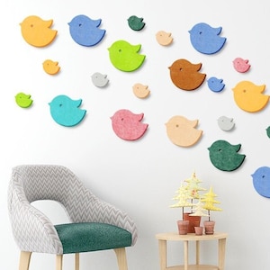 May include: A white wall adorned with a collection of felt bird wall decorations in various colours, including blue, green, yellow, orange, pink, and grey. The birds are arranged in a curved pattern, with the larger birds at the top and the smaller birds at the bottom. A green chair and a wooden table with two small potted trees are in the foreground.