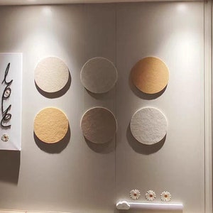 Può includere: Sei pezzi rotondi di arredamento da parete in feltro in varie tonalità di beige e marrone. I pezzi sono disposti in fila su una parete bianca.