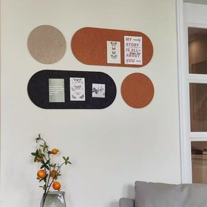May include: A white wall with four cork boards in different shapes and sizes. The boards are decorated with photos, postcards, and other ephemera. The boards are hung on the wall in a staggered pattern. There is a vase with orange flowers on a table in the foreground. The text on the postcard reads "My story is all about you."