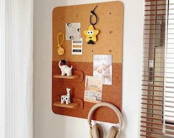 May include: A rectangular cork wall organizer board with two small wooden shelves, featuring a light brown upper section and a darker brown lower section. It holds a yellow star charm, a white elephant figurine, a giraffe figurine, photos, and beige headphones.