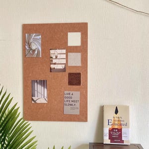 Puede incluir: Un corcho marrón con varias fotos pequeñas y muestras de color, junto con un pequeño letrero que dice "LIVE A GOOD LIFE MEET SLOWLY". Un libro titulado "Educated" está sobre una mesa auxiliar de madera.