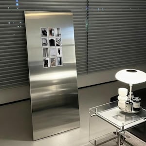 Puede incluir: Un espejo de pie rectangular y alto hecho de acero inoxidable cepillado con una superficie lisa y reflectante. Una cuadrícula de doce pequeñas impresiones fotográficas cuadradas en blanco y negro está fijada al centro del panel metálico.