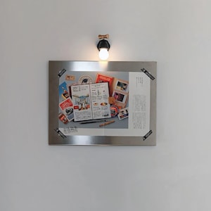 Puede incluir: Un marco met&aacute;lico rectangular muestra un collage de objetos relacionados con viajes, incluyendo un men&uacute;, fotograf&iacute;as y sellos. El marco est&aacute; montado en una pared bajo una peque&ntilde;a l&aacute;mpara de pared redonda con una bombilla c&aacute;lida.