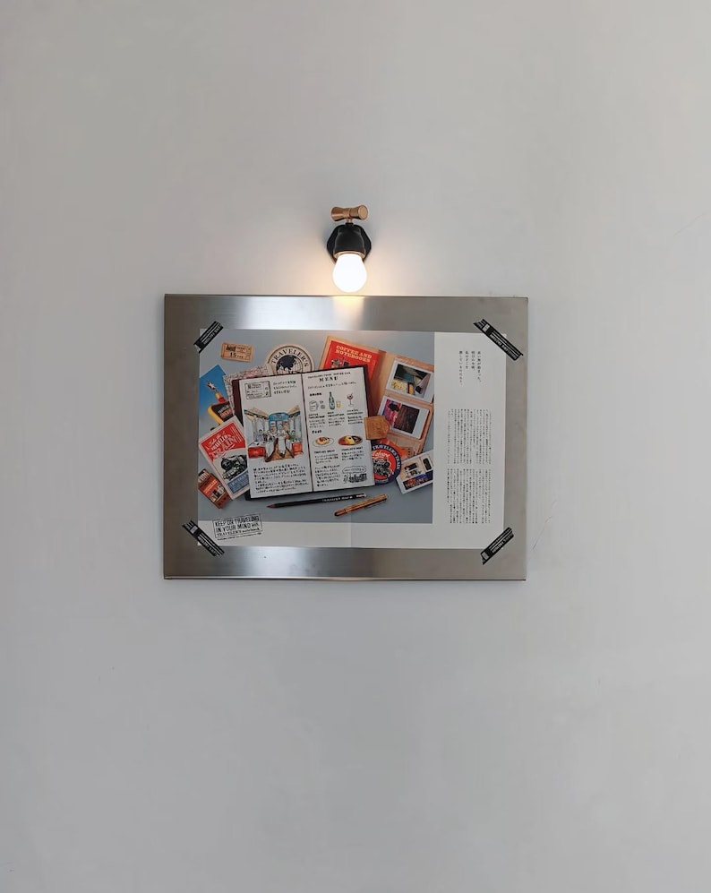 Puede incluir: Un marco met&aacute;lico rectangular muestra un collage de objetos relacionados con viajes, incluyendo un men&uacute;, fotograf&iacute;as y sellos. El marco est&aacute; montado en una pared bajo una peque&ntilde;a l&aacute;mpara de pared redonda con una bombilla c&aacute;lida.
