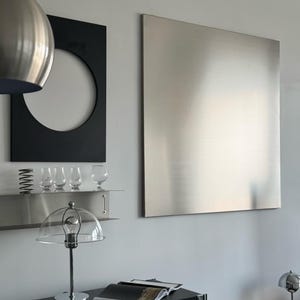 Puede incluir: Un gran panel de pared cuadrado de metal cepillado cuelga junto a una pieza de arte mural rectangular negra con un recorte circular. Debajo, un estante de metal sostiene cuatro vasos de vidrio transparente peque&ntilde;os y un resorte de metal.