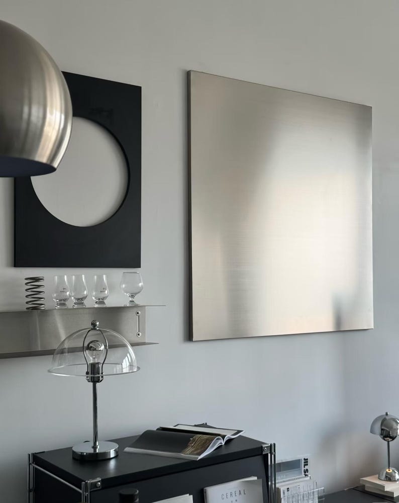 Puede incluir: Un gran panel de pared cuadrado de metal cepillado cuelga junto a una pieza de arte mural rectangular negra con un recorte circular. Debajo, un estante de metal sostiene cuatro vasos de vidrio transparente peque&ntilde;os y un resorte de metal.