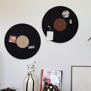 May include: Two black round cork boards shaped like vinyl records with cork inserts in the center. The boards are mounted on a white wall. The cork boards are decorated with various items, including photos, a small camera, and a tag.