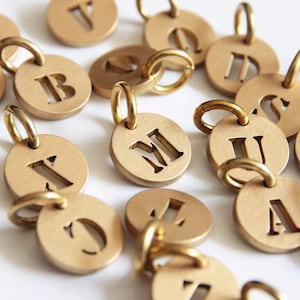 May include: Gold-colored metal alphabet charms with a circular design. Each charm features a different letter of the alphabet cut out in the center and a small loop at the top for attaching to a chain or bracelet.