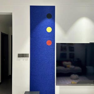 May include: A rectangular, deep blue felt wall panel with three colored circles: black, yellow, and red. The panel is mounted on a white wall, with a black side table and a television visible in the background. The room is well-lit.