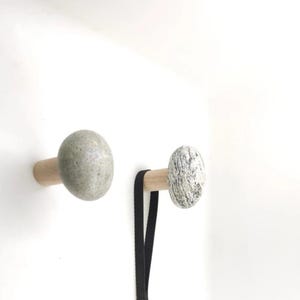 May include: Two wall-mounted coat hooks. Each hook has a light-coloured wooden base with a rounded, stone-like knob. One knob is speckled grey, the other is a solid grey. A black lanyard hangs from one hook.
