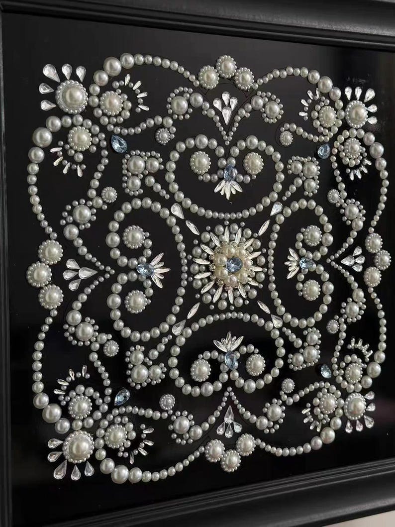 Op de afbeelding: Een ingelijst kunstwerk met een symmetrisch ontwerp van witte imitatieparels en heldere kristallen op een zwarte achtergrond. Het ontwerp bevat bloemen- en geometrische patronen, met accenten van lichtblauwe kristallen. De lijst is zwart.
