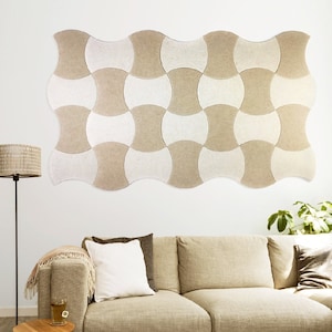 Puede incluir: Un panel acústico de fieltro beige y blanco en forma de onda, montado en una pared blanca sobre un sofá beige con cojines.