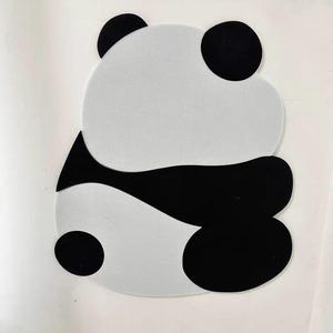 May include: A black and white felt panda wall decoration. The panda is in a sitting position with its arms crossed. The panda is made of two pieces of felt, one black and one white.