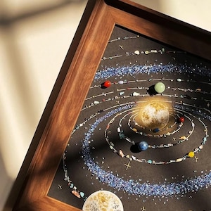 May include: A framed artwork depicting a solar system. The planets are represented by colorful gemstones and beads arranged on a black background. The frame is made of dark wood, and the artwork is lit by a warm light source.