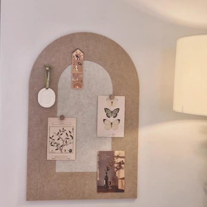 May include: A beige felt memo board with an arched top, displaying botanical prints and a decorative tag. The board features a light grey centre section and is adorned with small wooden pins. A white wax seal and a decorative bookmark are also attached.