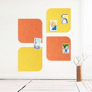 May include: Four square, felt wall decor pieces in yellow and orange are mounted on a white wall. Small pictures and stickers are attached to the felt squares. A brown vase with branches is in the lower right corner.