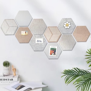 Può includere: Una collezione di pannelli murali esagonali in feltro in vari colori neutri, tra cui beige, grigio e bianco. Alcuni pannelli presentano motivi geometrici o piccole carte con immagini e testo. Una margherita di legno e il testo "Have a nice day" sono visibili.