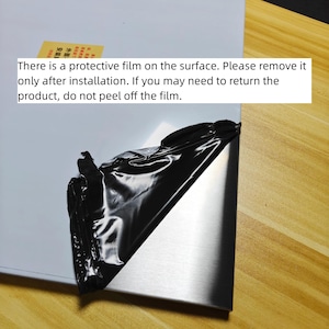 Puede incluir: Un panel met&aacute;lico rectangular con acabado plateado cepillado, parcialmente cubierto por una pel&iacute;cula protectora negra que se est&aacute; despegando. Una pegatina amarilla con texto en chino es visible en la superficie blanca del panel.