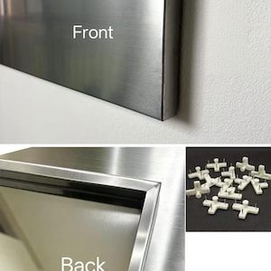 Puede incluir: Un panel met&aacute;lico rectangular con acabado cepillado, etiquetado como Front y Back en las im&aacute;genes. La vista trasera muestra un borde de marco empotrado. Se incluye un grupo separado de peque&ntilde;os colgadores de pared de pl&aacute;stico blanco con pines.