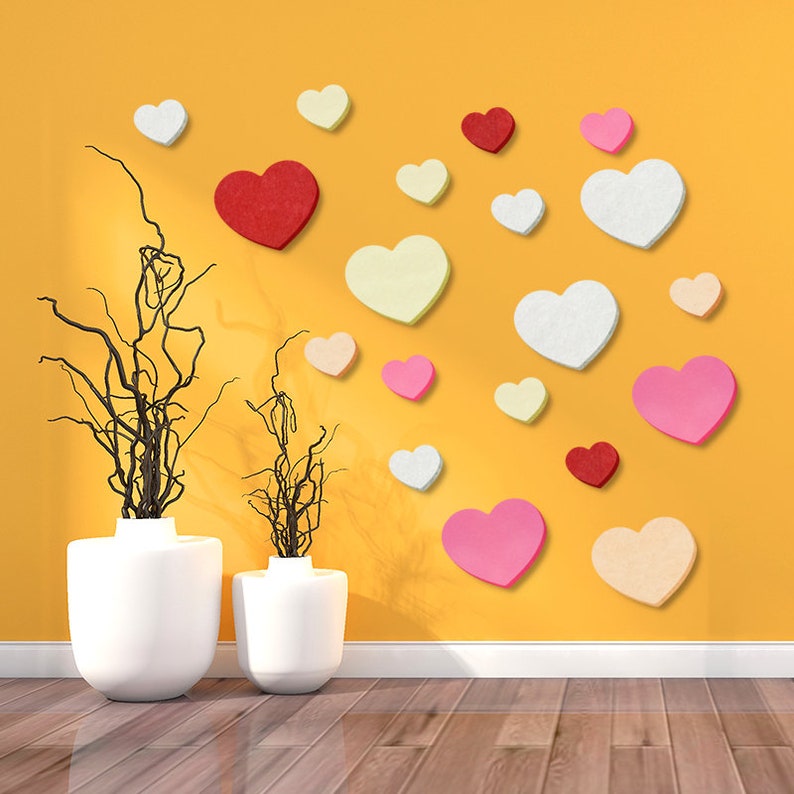 May include: A yellow wall decorated with felt hearts in various sizes and colors, including red, pink, white, and cream. Two white ceramic vases with brown branches are in the foreground.