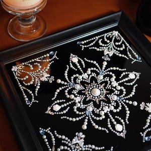 Op de afbeelding: Een vierkante, zwarte decoratieve tray met een ingewikkeld bloemenontwerp. Het ontwerp is gemaakt van witte parels, heldere kristallen en kleine blauwe accenten. De tray is omlijst door een zwarte houten rand, wat een opvallend contrast cre&euml;ert.