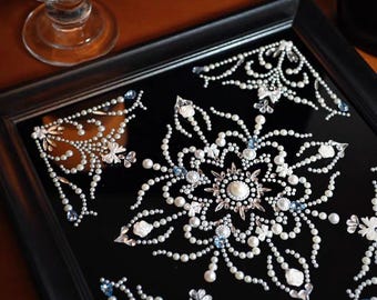 May include: A square, black decorative tray with an intricate floral design. The design is made of white pearls, clear crystals, and small blue accents. The tray is framed in a black wooden border, creating a striking contrast.