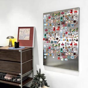 Puede incluir: Un gran tablero magn&eacute;tico rectangular de acero inoxidable montado en una pared blanca, densamente cubierto con numerosos pines e imanes coloridos de diversas formas, tama&ntilde;os y dise&ntilde;os, dispuestos en un patr&oacute;n de cuadr&iacute;cula en la superficie.