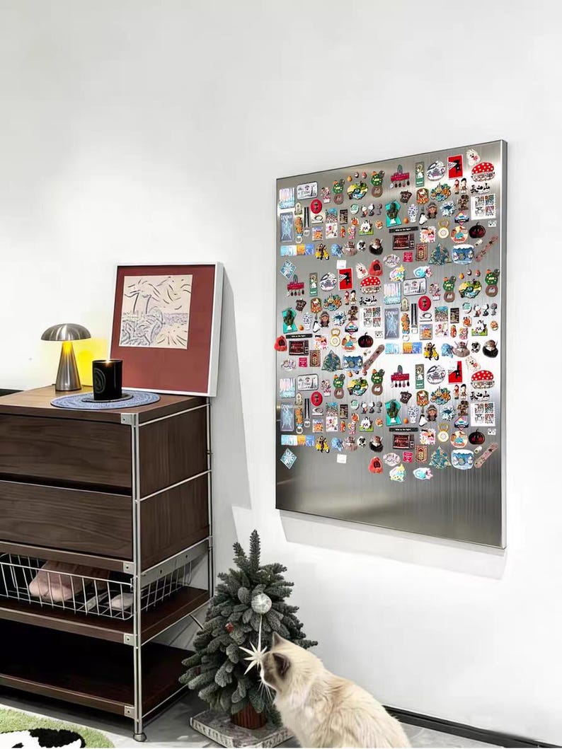 Puede incluir: Un gran tablero magn&eacute;tico rectangular de acero inoxidable montado en una pared blanca, densamente cubierto con numerosos pines e imanes coloridos de diversas formas, tama&ntilde;os y dise&ntilde;os, dispuestos en un patr&oacute;n de cuadr&iacute;cula en la superficie.