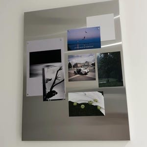 Puede incluir: Un tablero magn&eacute;tico rectangular de metal plateado que muestra varias fotograf&iacute;as y una tarjeta blanca en blanco. Las im&aacute;genes incluyen un atardecer con p&aacute;jaros, un coche en una carretera, un bosque oscuro, una silueta de &aacute;rbol en blanco y negro y manzanas verdes.