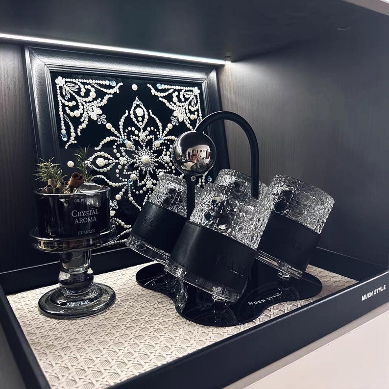 Op de afbeelding: Een decoratieve display met een zwart omlijst kunstwerk met een wit kralenontwerp, een kleine potplant en een set van drie kristallen glazen met zwarte leren banden. De display staat op een geweven mat.