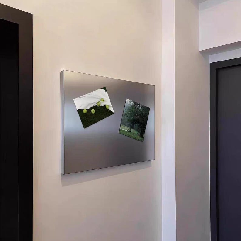 Puede incluir: Un tablero magn&eacute;tico rectangular de tono plateado montado en una pared. El tablero presenta una superficie met&aacute;lica lisa y reflectante con dos impresiones rectangulares adheridas, una que muestra manzanas verdes y otra un parque.