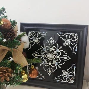 Op de afbeelding: Een ingelijst kunstwerk met een gedetailleerd sneeuwvlokontwerp. Het kunstwerk heeft witte en zilveren versieringen op een zwarte achtergrond, in een zwart frame. Een kleine kerstboom met ornamenten staat op de voorgrond.