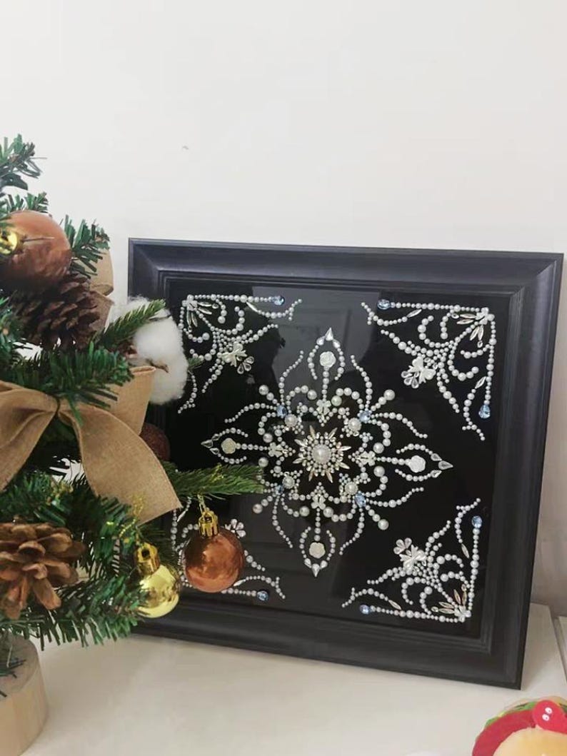 Op de afbeelding: Een ingelijst kunstwerk met een gedetailleerd sneeuwvlokontwerp. Het kunstwerk heeft witte en zilveren versieringen op een zwarte achtergrond, in een zwart frame. Een kleine kerstboom met ornamenten staat op de voorgrond.