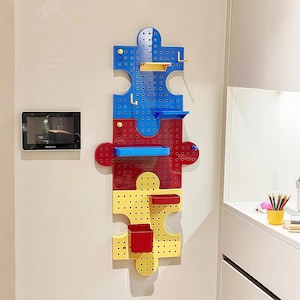 Può includere: Un colorato organizer da parete a forma di pezzo di puzzle. L'organizer è composto da sezioni blu, rosse e gialle, ciascuna con vari ripiani e scomparti. Include piccoli ganci e contenitori per riporre oggetti.