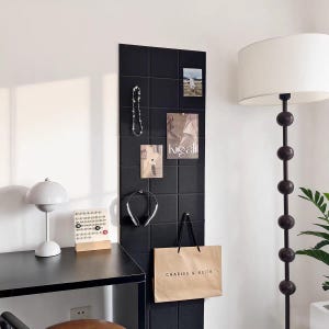 Puede incluir: Un organizador de pared negro con azulejos cuadrados con varios elementos adjuntos, incluyendo un collar, fotos, unos auriculares plateados y una bolsa de compras de Charles & Keith. Una lámpara de escritorio blanca y un calendario están a la izquierda.