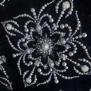 Op de afbeelding: Symmetrisch decor op een zwarte ondergrond. Het bestaat uit witte parels, heldere kristallen en kleine witte rozen- en strikaccenten. Lichtblauwe edelstenen zijn in het ontwerp verwerkt.