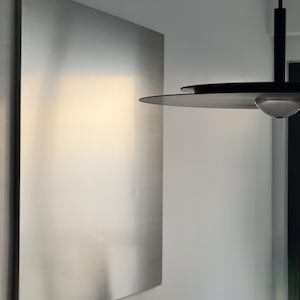 Puede incluir: Un panel de pared rectangular grande de metal plateado cepillado con una superficie lisa y reflectante. Al lado cuelga una l&aacute;mpara colgante negra con una pantalla circular plana y delgada y una peque&ntilde;a bombilla de cristal transparente y redondeada en el centro.