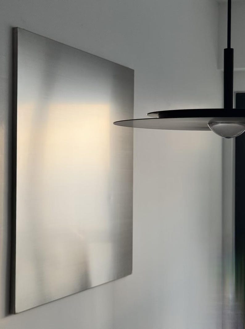 Puede incluir: Un panel de pared rectangular grande de metal plateado cepillado con una superficie lisa y reflectante. Al lado cuelga una l&aacute;mpara colgante negra con una pantalla circular plana y delgada y una peque&ntilde;a bombilla de cristal transparente y redondeada en el centro.