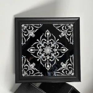 Op de afbeelding: Zwart omlijst kunstwerk met een symmetrisch ontwerp van witte parels en kristallen op een zwarte achtergrond. Het ontwerp is een bloemachtig mandala-achtig patroon. De lijst is vierkant en zwart.