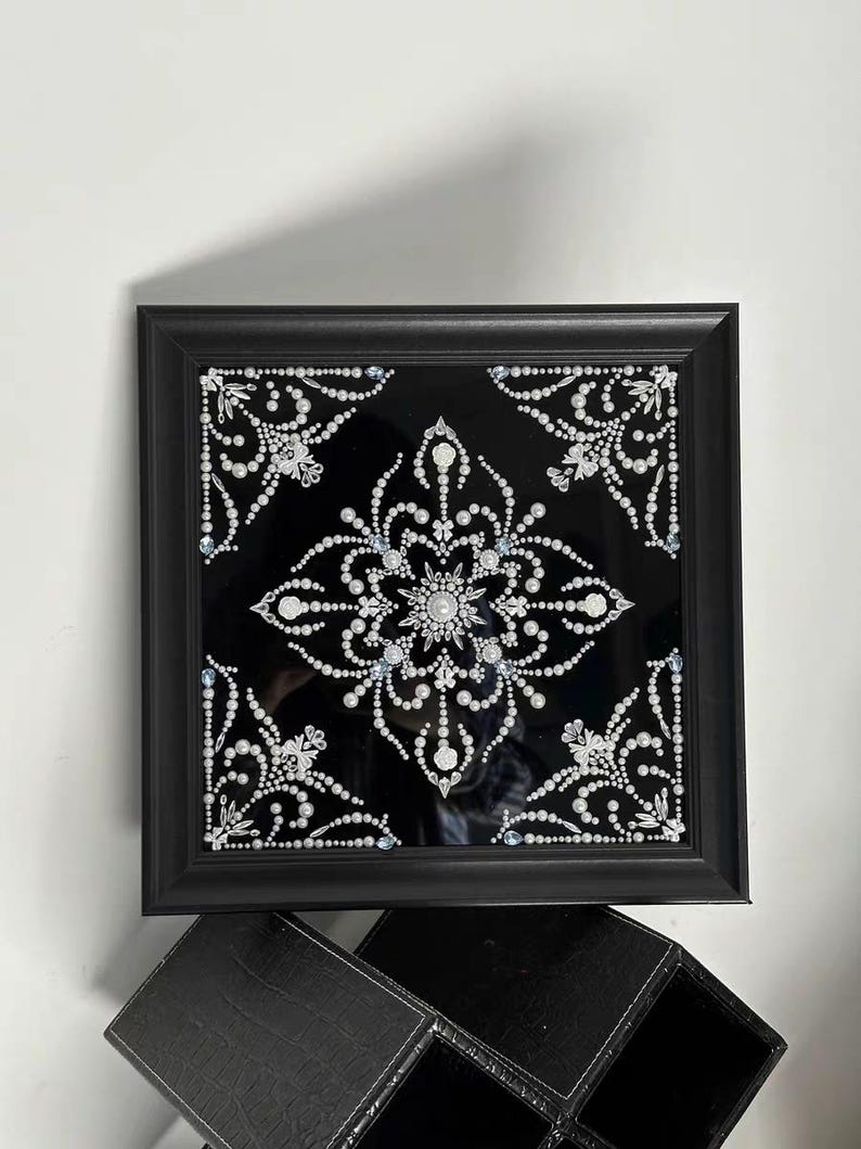 Op de afbeelding: Zwart omlijst kunstwerk met een symmetrisch ontwerp van witte parels en kristallen op een zwarte achtergrond. Het ontwerp is een bloemachtig mandala-achtig patroon. De lijst is vierkant en zwart.