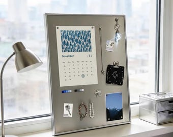 Puede incluir: Un tablero de notas magnético rectangular de color plateado que muestra una página de calendario de noviembre con un patrón de pinos azules, un clip cuadrado negro, una foto de paisaje de montaña, un imán de estrella, joyas y notas.
