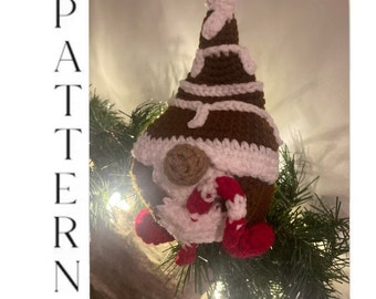 May include: A crocheted gingerbread gnome ornament with a white hat and red candy cane. The gnome is decorated with white icing and brown details. The ornament is hanging on a green Christmas tree branch.